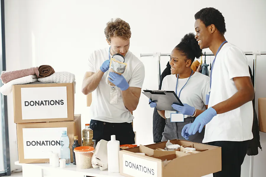 Three volunteers in gloves checking donated groceries against a clipboard beside labelled donation boxes